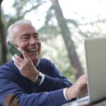 Happy senior man talking on phone and using laptop outdoors.